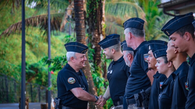 Le DGGN, en polo noir et pantalon de treillis, calot sur la tête, serre la main d'un gendarme dans la même tenue. D'autres gendarmes sont en rang à côté de lui, au garde-à-vous.