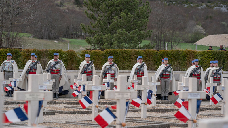 Agrandir l'image Image d'un cimetière aux croix blanches, sur lesquelles sont disposés des drapeaux français. Des militaires se tiennent debout en bordure des tombes.