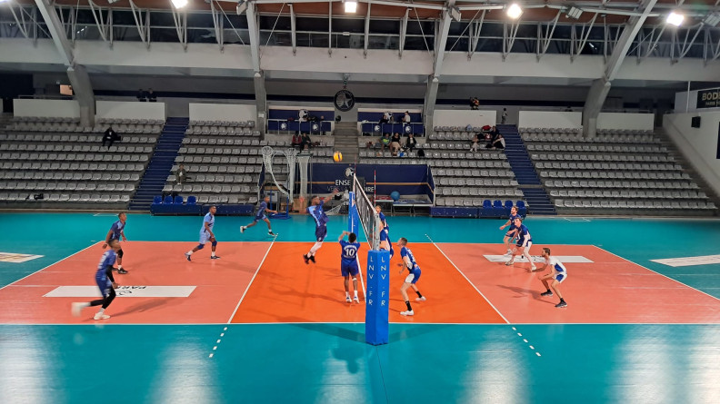 Dans un gymanse au sol bleu et orange, dexu équipes s'affrontent au voleyball