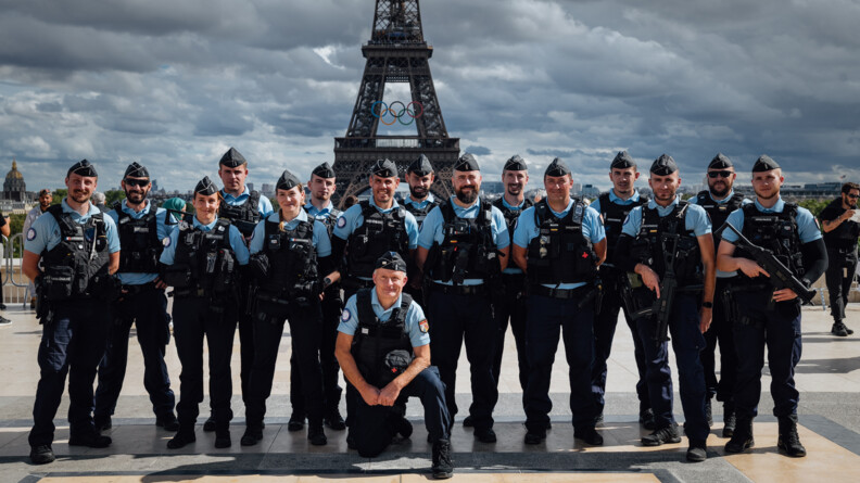Des gendarmes posent devant et sur la Place du Trocadéro, à Paris. Derrière eux se dresse la tour Eiffel, sur laquelle sont aposés les anneaux des Jeux olympiques et paralympiques.