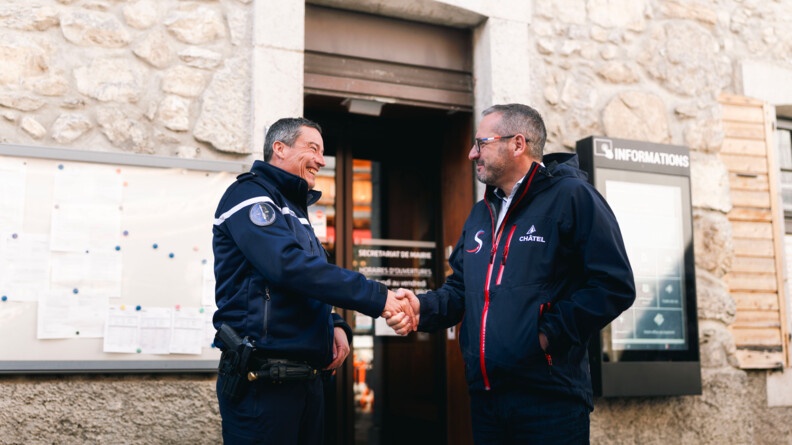 Agrandir l'image Un gendarme serre la main du maire de la commune de Châtel. Les deux hommes se tiennent debout, devant la façade de la mairie. Ils se sourient.