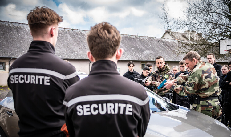 Agrandir l'image Cours d'intervention professionnelle au profit des lycéens - au premier plan vers la gauche, deux jeunes hommes de dos vêtus d'un pull noir floqué "sécurité" en blanc. Au second plan, derrière une voiture Renault grise, deux militaires effectuent une démonstration d'un contrôle avec arme factice. Ils sont entourés d'une dizaine de jeunes qui suivent la démonstration. En arrière-plan, un bâtiment en longueur recouvert d'ardoises.