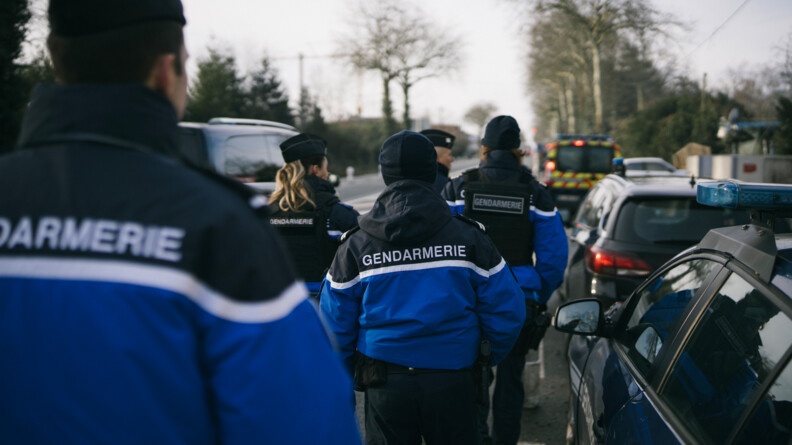Agrandir l'image 5 gendarmes de dos dans une rue. Sur les côtés quelques voitures garées.