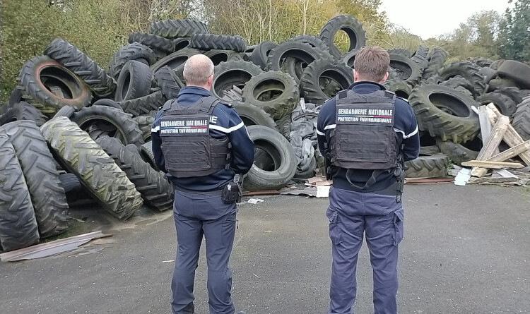 Agrandir l'image Deux gendarmes de dos, devant un monticule de pneus de camion et de tracteur posés sur le bitume, à la lisière d'un bois.