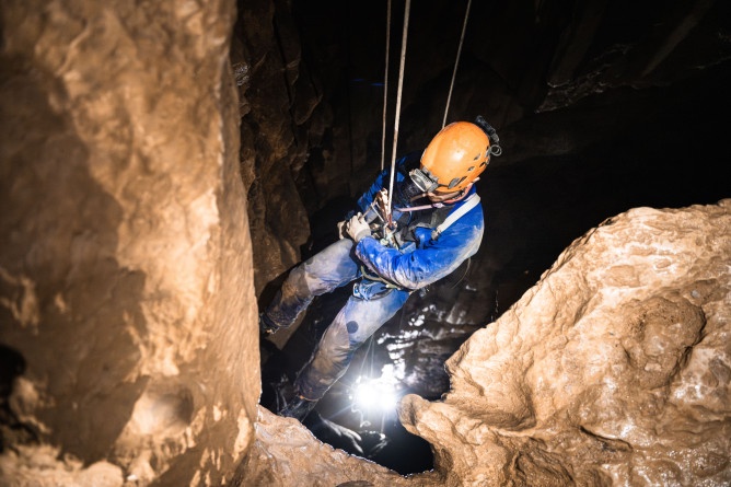 Agrandir l'image Photo montrant un gendarme spéléologue en combinaison et casque équipé d’une lampe frontale, en train de descendre le long d'une paroi rocheuse à l’intérieur d’une grotte.