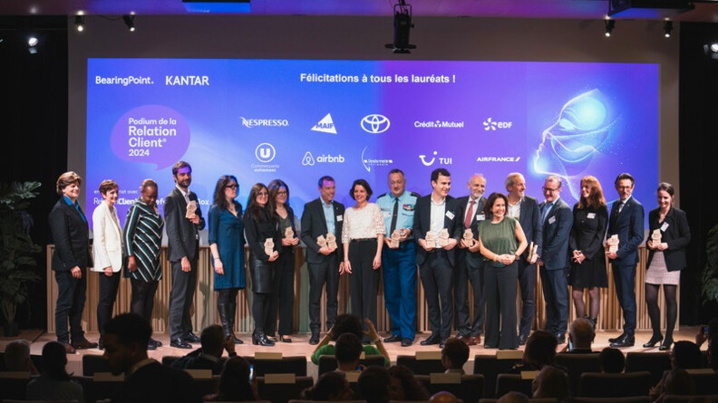 Dix-huit personnes, hommes et femmes, sur une sc&egrave;ne, tenant leurs troph&eacute;es de la Relation client 2024. Au centre, le directeur g&eacute;n&eacute;ral de la gendarmerie, en tenue bleue. En fond, un &eacute;cran bleu/violet, o&ugrave; sont projet&eacute;s les logos des laur&eacute;ats 2024.