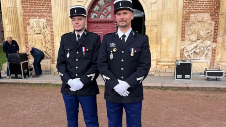 Agrandir l'image Deux gendarmes en uniforme posent côte à côte devant un édifice en pierres blanches et rouges.