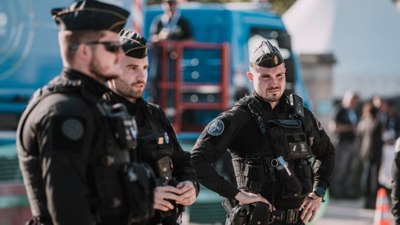 Trois gendarmes mobiles de face et de profil et en fond, flou, un camion régie de la cérémonie d'Omaha Beach.