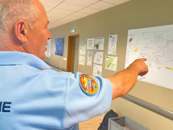 Agrandir l'image Un gendarme en polot manches courtes, vu de trois quarts dos, pointe du doigt une carte accrochée sur un mur.