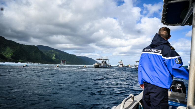 Un gendarme de dos sur une embarcation qu'on aperçoit le bord à droite de l'image. Sur le plan d'eau, des bateaux et jet-ski devant des montagnes vertes de Tahiti
