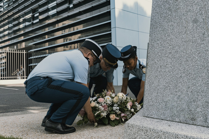 Trois gendarmes d&eacute;posent ensemble une gerbe de fleurs au pied d'un monument, dans une cour. La sc&egrave;ne se d&eacute;roule de jour.