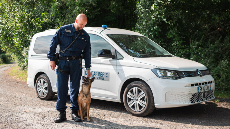Un maître de chien de plein-pied, en combinaison bleue-marine, avec un malinois assis à ses pieds qui le regarde. Au fond le véhicule blanc de l'équipe