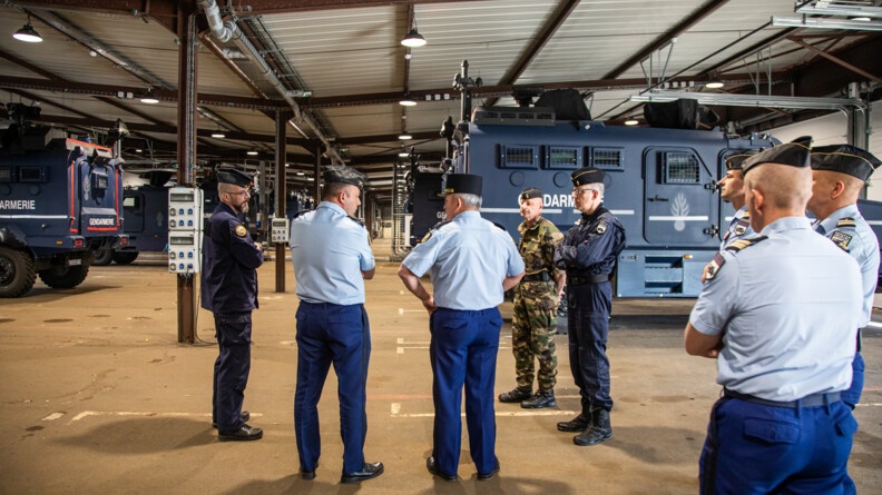 Agrandir l'image Le directeur général de la gendarmerie et le général commandant le GBGM vus de dos, dans un hangar. Devant eux, plusieurs véhicules Centaure. Ils sont entourés de plusieurs gendarmes en tenue, trois en bleu et deux en kaki.