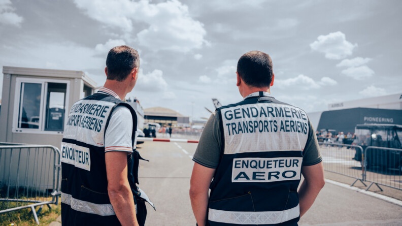 De dos, face à une barrière rouge et blanche, deux enquêteurs de la GTA en civil, avec une chasuble bleu marine réhaussée de blanc, indiquant "enquêteur" (gendarme de gauche) et "enquêteur aéro" (gendarme de droite)