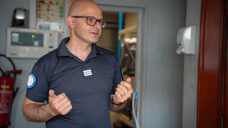 Un homme d'âge moyen se tient debout, face à l'objectif, dans une pièce, derrière un comptoir. Il discute, s'animant avec les mains. Il porte un t-shirt bleu marine et des lunettes noires. L'homme est chauve.