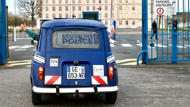 Une Renaud 4L bleu marine p&eacute;n&egrave;tre dans l'enceinte d'un grand b&acirc;timent que l'on aper&ccedil;oit en fond. On voit l'arri&egrave;re de la voiture, portant sur la vitre arri&egrave;re la mention "Ecussons ploice.com". On aper&ccedil;oit, sortant par la fen&ecirc;tre, le pouce tendu en signe de victoire, du conducteur et de son passager.
