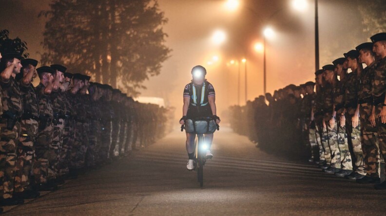 Agrandir l'image Une jeune femme cycliste remonte une allée de nuit, tout au long de laquelle se dresse une haie d'honneur de militaires en tenue. Les lamapadaires et la lampe frontale de la cycliste forment des points lumineux.