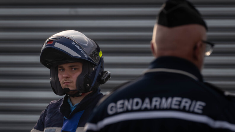 un motard de la gendarmerie en buste avec son casque relevé. Devnat lui, flou, à droite, un autre gendarme