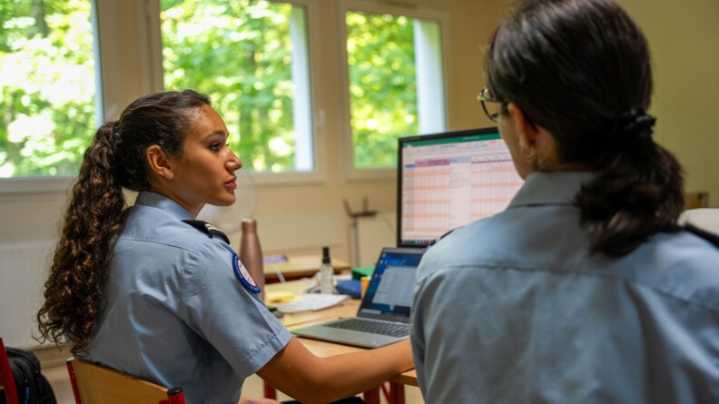 Deux femmes gendarmes en uniforme, sont assises côte à côte, dans une salle, devant une table sur laquelle sont posés plusieurs ordinateurs. On aperçoit les arbres par la fenêtre