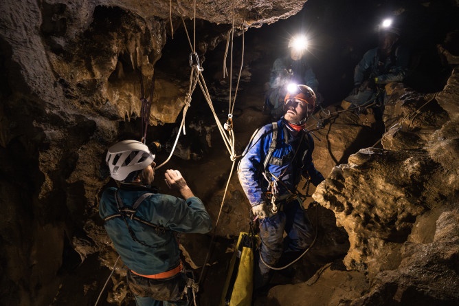 Agrandir l'image Photo montrant plusieurs gendarmes spéléologues équipés de combinaisons, de casques et de lampes frontales, évoluant dans une grotte. Ils sont attachés à des cordes et utilisent du matériel d’escalade pour progresser parmi les parois rocheuses et les formations calcaires.