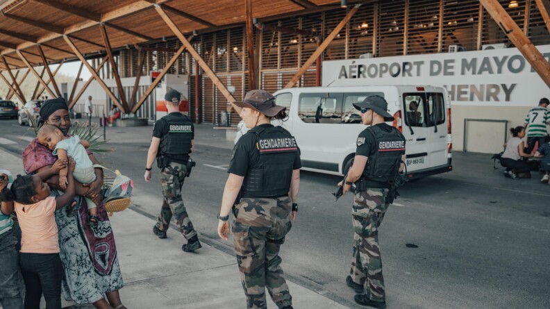 trois gendarmes en treillis et haut noir patrouilles, &agrave; gauche une femme avec ses enfants, &agrave; droite un v&eacute;hicule blanc gar&eacute; devant l'a&eacute;roport de mayotte