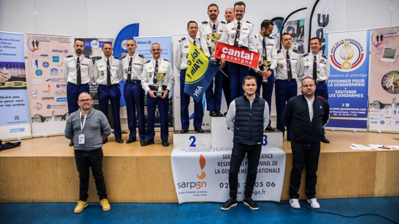Agrandir l'image 11 coureurs en tenue chemise blanche avec leurs médailles au cou sur une scène et le podium; en fond des kakémonos gendarmerie et sponsors; au pied de la scène trois hommes en civils mains derrière le dos