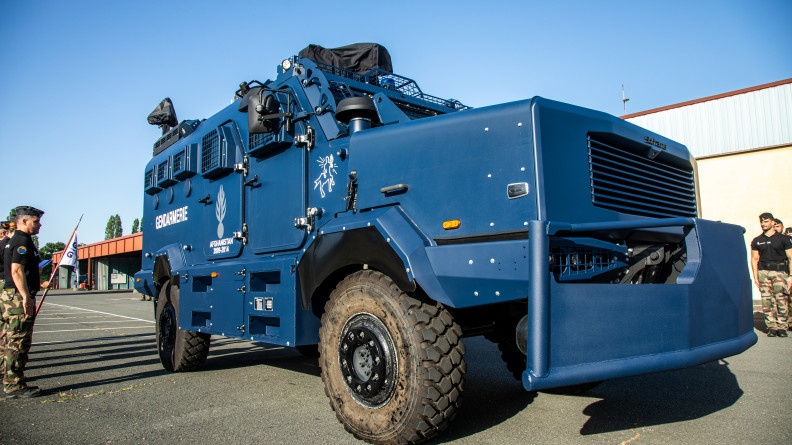 Agrandir l'image La photo représente un véhicule blindé de la gendarmerie avançant devant des mililtaires alignés au bord de la route