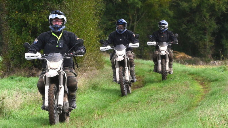 Trois moto cross conduites par des gendarmes en treillis et haut noir sur un chemin herbeux le long des bois