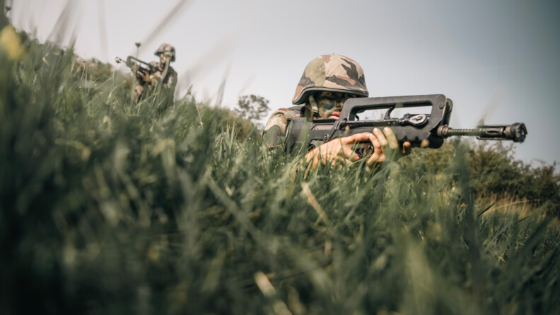 Militaire couch&eacute; dans l'herbe pointant son arme en direction de l'ennemi. Son chef de trin&ocirc;me se trouve &agrave; l'arri&egrave;re.
