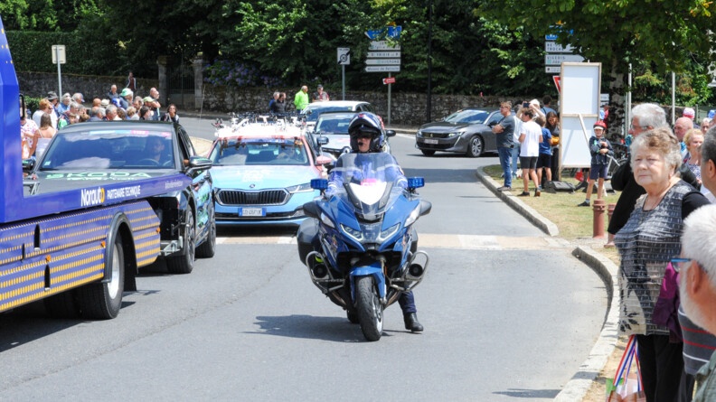 Agrandir l'image Un motocycliste gendarmerie prêt à mettre pied-à-terre au centre de l'image; de part et d'autre de la route des spectateurs ; à l'arrière de la moto plusieurs véhicules