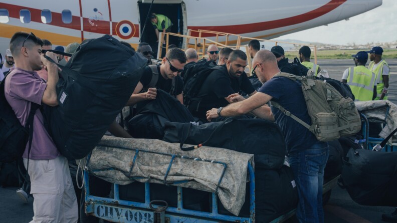 des gendarmes en civil chargent des sacs sur un chariot au pied d'un avion de la s&eacute;curit&eacute; civile