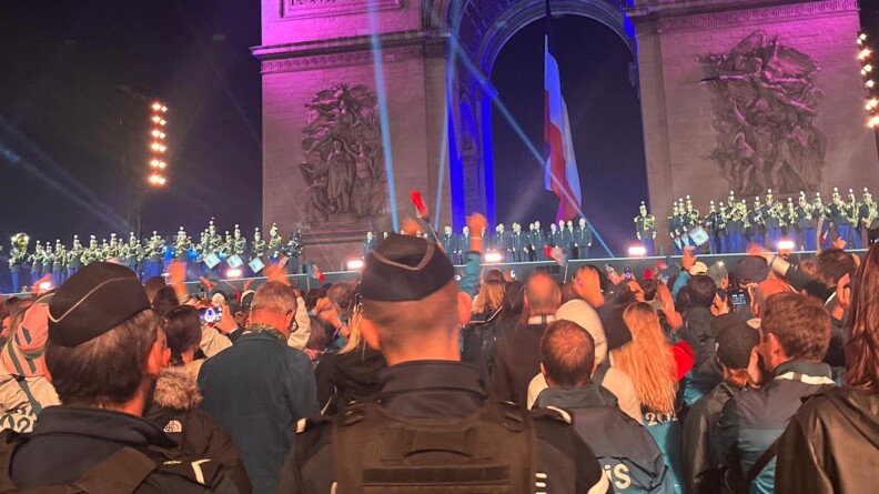 Sur une grande sc&egrave;ne install&eacute;e au pied de l'Arc de Triomphe, o&ugrave; flotte le drapeau fran&ccedil;ais, les musiciens de la Garde R&eacute;publicaine occupent les deux tiers de la sc&egrave;ne ; &agrave; droite, v&ecirc;tus de blanc, les athl&egrave;tes olympiques et paralympiques saluent la foule amass&eacute;e devant la sc&egrave;ne. Au premier plan, deux gendarmes d&eacute;partementaux, de dos, issus d'une des compagnies de marche mobilis&eacute;es pour assurer la s&eacute;curisation des festivit&eacute;s.