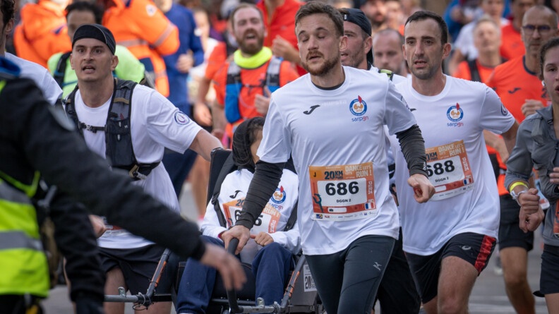 Une foule de coureurs, vêtus d'un T-shirt blanc avec un numéro sur le ventre, courent côte à côte. L'un d'eux pousse un fauteuil roulant.