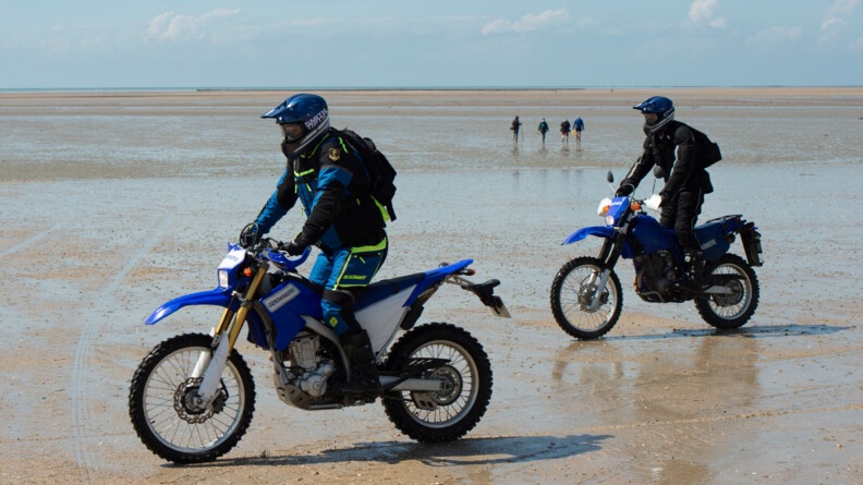 Deux motos cross de la gendarmerie sur une plage. Au fond quatre personnes de dos marchent la plage à marée basse
