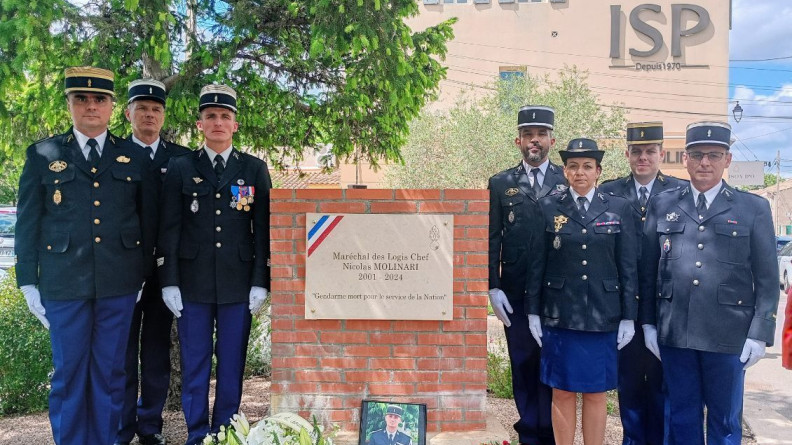 Sept gendarmes en vareuse debout de part et d'autre une plaque apposée sur une stèle en briques rouges, au pied de laquelle se trouvent une gerbe et le portrait d'un jeune gendarme décédé en Nouvelle-Calédonie.