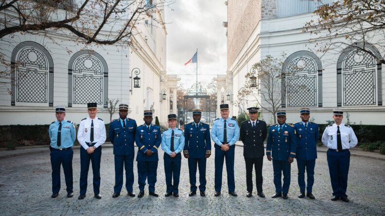 Dans une cour pavée se tiennent debout côte à côte onze hommes, de la gendarmerie française et de la gendarmerie congolaise. Derrière eux, se trouve les murs d'un bâtiment en pierre. Au centren on aperçoit un grand portail noir au dessus duquel flotte le drapeau français. Le ciel est nuageux.