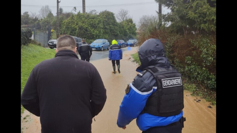 Trois gendarmes sous la pluie, &eacute;changent en marchant dans une rues inond&eacute;e avec un habitant.