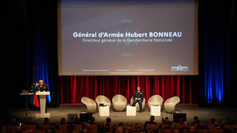 Sur une sc&egrave;ne, &agrave; gauche, le g&eacute;n&eacute;ral d'arm&eacute;e Bonneau, en vareuse, s'exprime au micro, derri&egrave;re un pupitre. A sa gauche, sur une table haute, un ordinateur est ouvert. Derri&egrave;re lui, les plis des drapeaux fran&ccedil;ais et europ&eacute;en. A sa droite, sur la sc&egrave;ne, le major g&eacute;n&eacute;ral, en vareuse, est assis sur un fauteuil blanc. Il est entour&eacute; de cinq autres fauteuils vides. En fond, sur un rideau rouge, un &eacute;cran g&eacute;ant indiquant "G&eacute;n&eacute;ral d'arm&eacute;e Hubert Bonneau, directeur g&eacute;n&eacute;ral de la gendarmerie nationale".