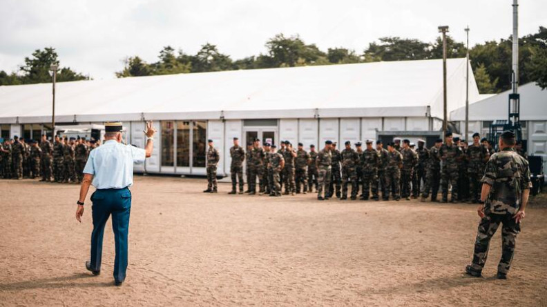 Agrandir l'image Un gendarme passe devant un groupe de militaires en tenue kakie, effectuant le signe de la victoire de sa main droite.