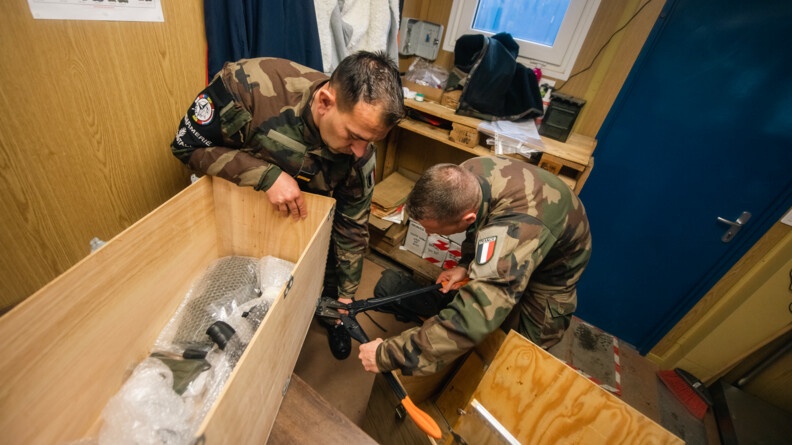 Agrandir l'image Vus d'en haut, un prévôt et un militaire préparent un colis sensible pour le vol, contenant des armes. Le colis, en bois, est à gauche de l'image, sur une table, et les militaires utilisent une grosse pince coupante