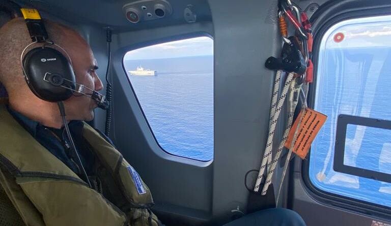 Vue sur la mer et le porte hélicoptères Mistral depuis le hublot d'un hélicoptère de la gendarmerie nationale. Au premier plan un militaire de l'équipage.