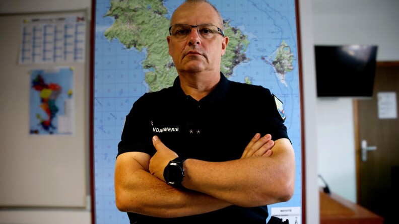 Photo en buste du général Lucien Barth, commandant la gendarmerie de Mayotte, les bras croisés devant lui, vêtu d'un tee-shirt noir portant la mention GENDARMERIE en blanc et deux étoiles au centre. En arrière-plan, sur le mur, une grande carte de Mayotte.