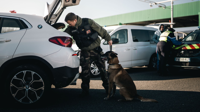 Agrandir l'image Image d'un malinois, photographié de biais, assis, aux côtés de son maître, un gendarme, fouillant le coffre d'un véhicule blanc. En arrière plan, on distingue la gare de péage de l'autoroute.
