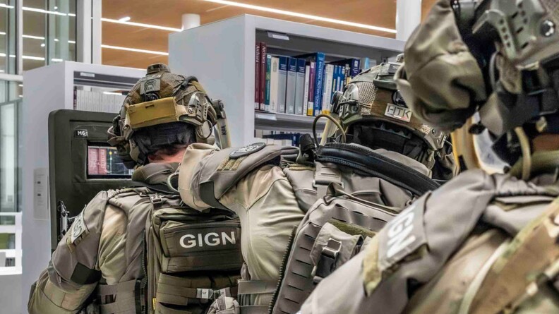 Trois équipiers de la FI progressent derrière un bouclier, dans les rayons de la bibliothèque.