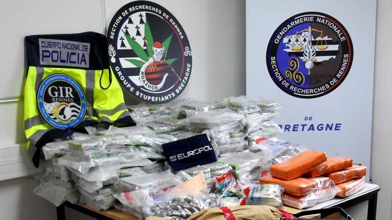 Des scellés sur une table avec en fond des kakemono de la Section de recherches de Rennes, du GIR de Rennes, du groupe stupéfiants de Bretagne et d'Europol.