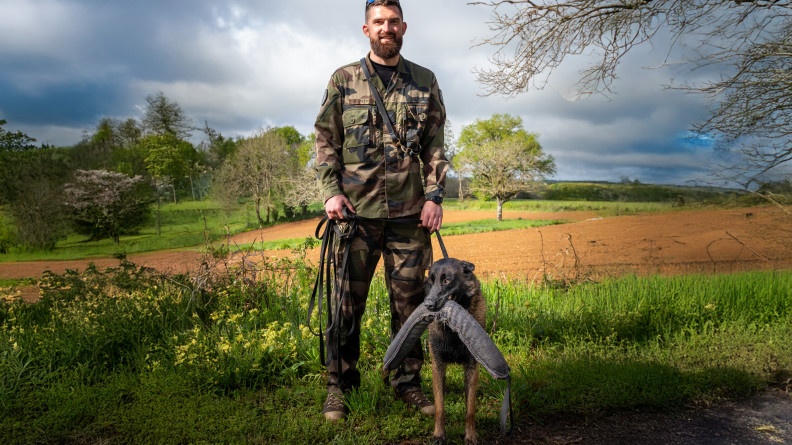 Photo d'un gendarme en tenu kakie, tenant en laisse en berger belge malinois, qui se tient &agrave; ses c&ocirc;t&eacute;s avec un jouet en tissu dans le gueule. La sc&egrave;ne se d&eacute;roule dans la campagne.