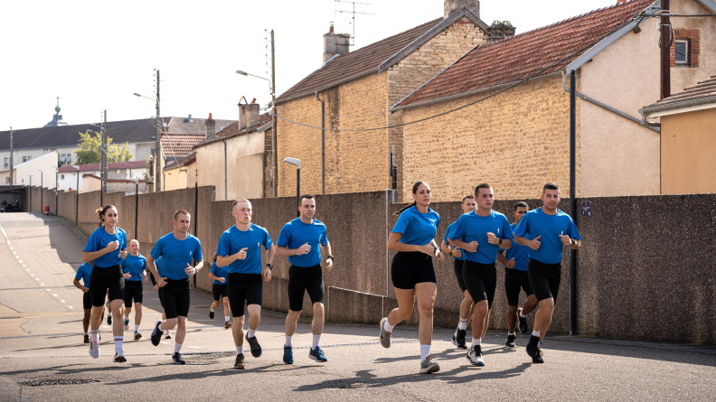 Une quinzaine d'élèves gendarmes en cyclistes noirs et t-shirts bleus en train de courir en groupe dans une enceinte militaire.