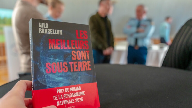 Une main gauche tient un livre dont le titre rouge "les meilleurs sont sous terreé se détache sur une couverture noire et bleue holographique. Une étiquette "prix du roman de la gendarmerie nationale 2025" recouvre le bas du livre. En fond, flou, des personnes discutent, dont un gendarme