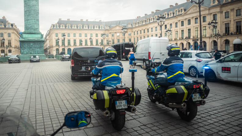 Deux gendarmes motocyclistes réalisant un eescorte arrivent Place Vendôme, à Paris, au guidon de leur véhicule. Nous les apercevons de dos. Autour d'eux se trouvent des voitures et des camionnettes.