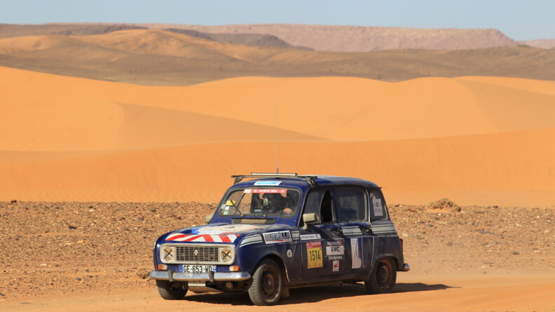 Une Renaud 4L bleu marine, couverte d'enseignes diverses, postionnées sur le flanc et le sur capot, roule en plein désert.
